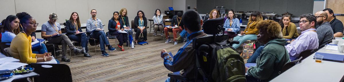 A group of approximately 30 people sitting on chairs in a circle while listening to the person in front speaking.